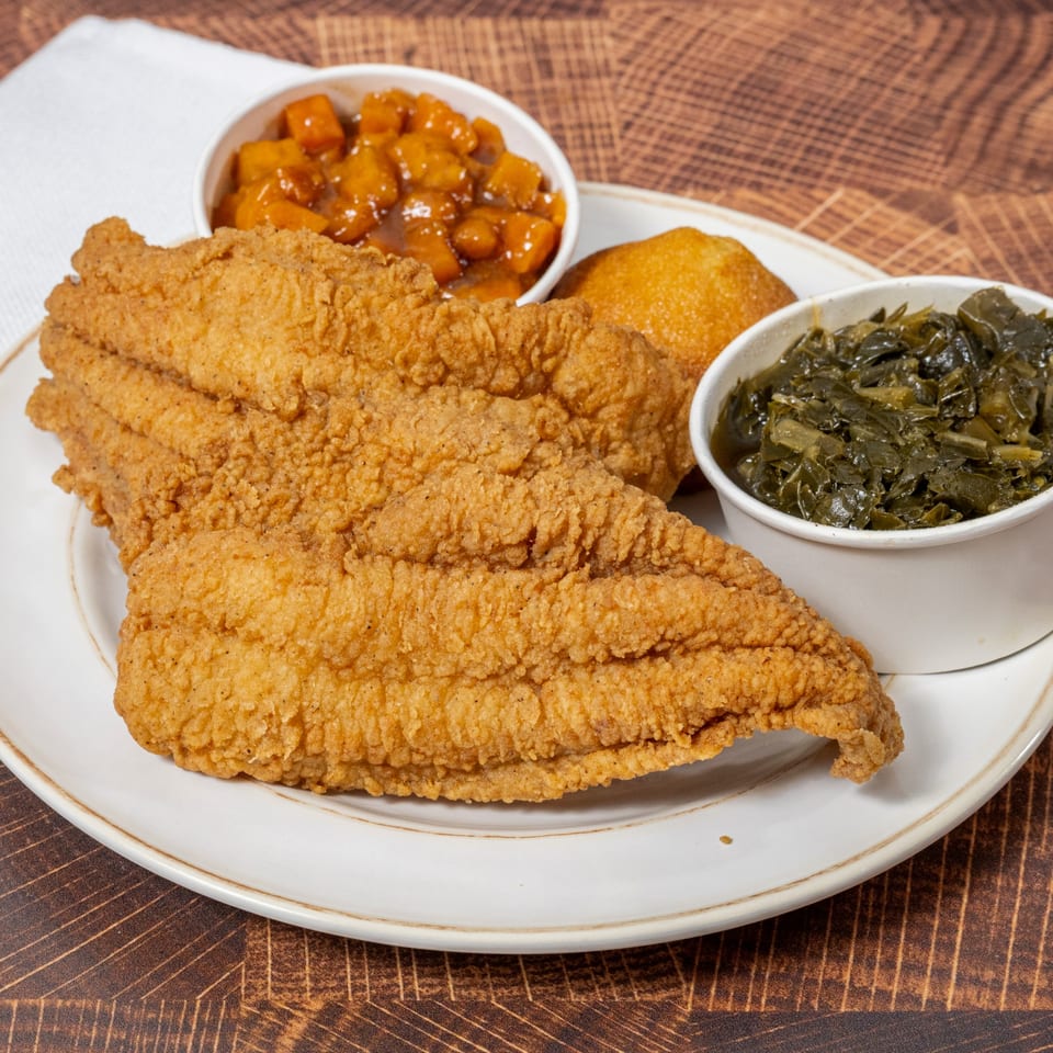 Fried fish fillets, candied yams, collard greens, and cornbread on a white plate from Chick N' Friends.